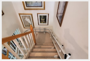 A carpeted staircase with a stairlift on the right side. The walls are adorned with framed art, including nature scenes and a bird. A wooden banister runs along the left side of the stairs.